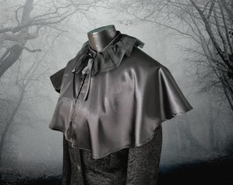 May include: A black satin cape with a ruffled collar and a tie at the neck. The cape is worn over a black patterned jacket.