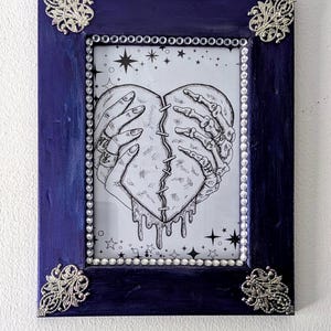 May include: A black and white illustration of a broken heart held together by a hand and a skeleton hand. The heart is dripping with a black substance. The illustration is framed in a dark blue frame with silver embellishments and rhinestones.