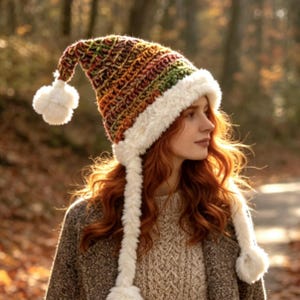 May include: A crochet hat featuring a variegated yarn pattern in shades of brown, green, and orange. The hat has a long, pointed shape with a white faux fur trim along the brim, two long braided side tassels, and three white pom-poms.