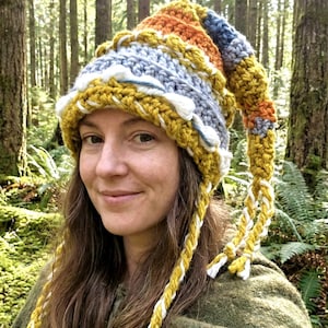 Puede incluir: Un sombrero de ganchillo hecho a mano y colorido con un extremo largo y puntiagudo. El sombrero presenta una mezcla de hilo amarillo mostaza, naranja, azul y gris, con detalles en blanco. Borlas trenzadas cuelgan de los lados. Este sombrero caprichoso es perfecto.