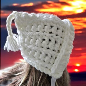Può includere: Un cappello bianco all'uncinetto fatto a mano con nappa e chiusura a fiocco. Il cappello presenta un motivo a punto strutturato e un bordo smerlato. Lo sfondo è un tramonto colorato.