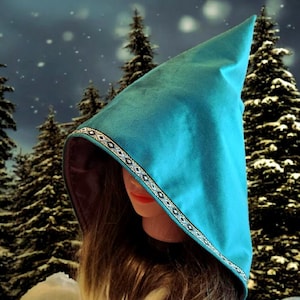 Puede incluir: Una capucha puntiaguda de color turquesa con un ribete decorativo. El interior de la capucha es de un tono más oscuro y está sobre un fondo de árboles de hoja perenne cubiertos de nieve. El ribete presenta un patrón geométrico en negro, blanco y plata.