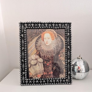 May include: A framed portrait of a woman with red hair and a ruffled collar. The black frame is decorated with small silver skulls and beads. A silver disco ball sits to the right of the frame.