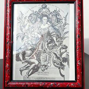 Op de afbeelding: Een rechthoekige fotolijst met een glanzende rode afwerking en een zwarte binnenrand, versierd met geschilderde zwarte ranken, kleine rode bloemen en witte schedels. Het bevat een zwart-wit historische prent met een koninklijk figuur en tekst.
