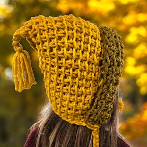 Può includere: Un cappello all'uncinetto giallo senape fatto a mano con una parte superiore a punta e un nappa. Il cappello ha un paraorecchie verde oliva a contrasto e un laccio. Il cappello è mostrato su uno sfondo sfocato di fogliame autunnale.
