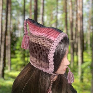 Puede incluir: Un gorro de punto de rayas rosas y marrones con una borla larga. El gorro tiene una corona puntiaguda y un ala ancha.