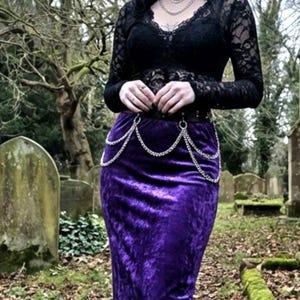 Op de afbeelding: Een zwarte kanten top met lange mouwen en een dieppaarse fluwelen rok. De rok is getailleerd en heeft een zilveren kettingriem. De outfit is gestyled in een gotische esthetiek.