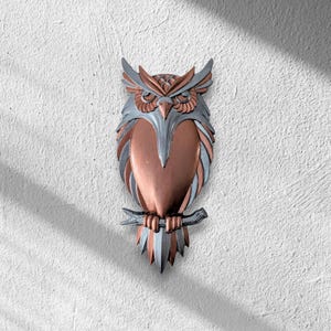 May include: A decorative owl wall hanging with a copper and silver finish. The owl is perched on a branch and has detailed feather and eye features. The wall art is mounted on a textured white wall.