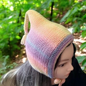 Puede incluir: Un gorro de crochet de color arcoíris pastel con una parte superior larga y puntiaguda y un borla. El gorro está hecho con un hilo suave y tiene una sensación cálida y acogedora.