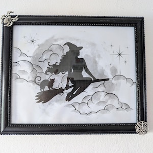 May include: Black framed art print featuring a silhouette of a witch riding a broom with a cat, set against a cloudy sky with stars. The frame has decorative corners with spider and web accents. A Halloween-themed wall decor.