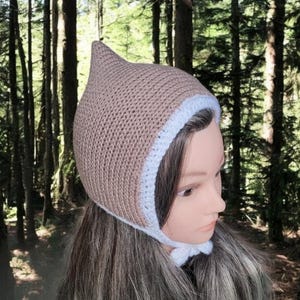 Puede incluir: Un gorro de bebé de ganchillo marrón con un borde de ganchillo blanco. El gorro tiene una parte superior puntiaguda y se ata debajo de la barbilla.