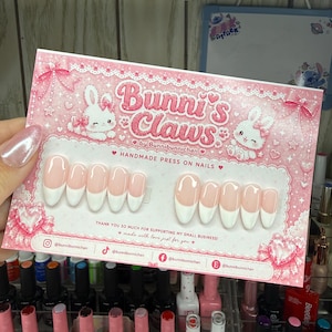 May include: A set of press-on nails with a French manicure design, displayed on a pink card with the text "Bunni's Claws." The card features illustrations of bunnies and hearts. The nails are pink with white tips. A variety of nail polish bottles are in the background.