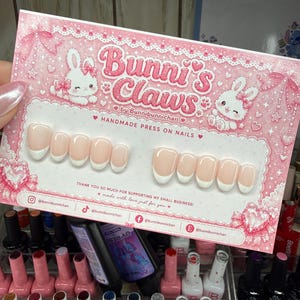 May include: A set of handmade press-on nails from Bunni's Claws, featuring a French tip design. The nails are displayed on a pink card with bunny illustrations and the text "Bunni's Claws". Various nail polish bottles are visible in the background.