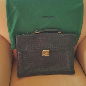 May include: A black leather briefcase with a woven pattern and gold hardware. The briefcase is sitting on a tan couch with a green dust bag that says "Bottega Veneta" on it.