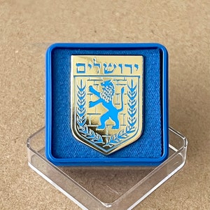 Puede incluir: Pin metálico dorado con un emblema de león azul y la palabra hebrea "Jerusalén". El pin está colocado en una caja forrada de terciopelo azul, exhibido sobre un soporte acrílico transparente.