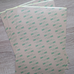 May include: A stack of 3M 467MP adhesive transfer tape sheets. The backing is a light tan colour, printed with a repeating green 3M logo and product details. The sheets are on a light brown surface.