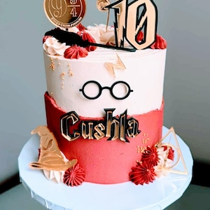 May include: A two-tiered cake with white and red frosting, decorated with gold Harry Potter-themed toppers. The cake features a Platform 9 3/4 sign, a golden snitch, glasses, a lightning bolt, and the name "Cushla".