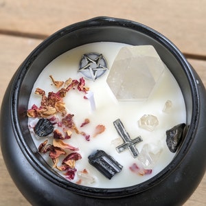May include: Black cauldron candle with a white wax top. The candle is decorated with dried rose petals, black crystals, and silver pentagram and cross charms.