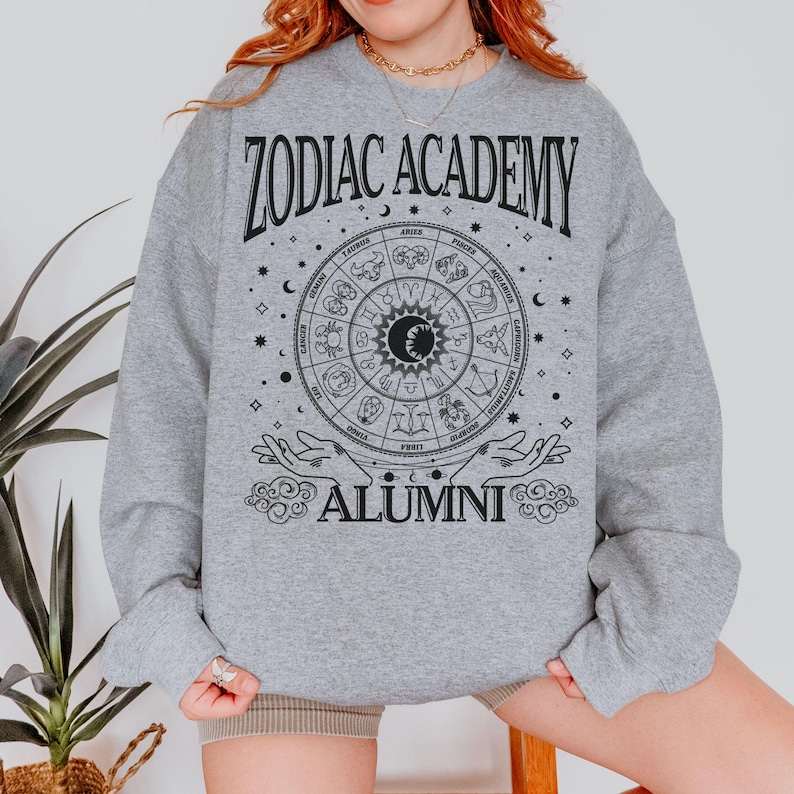 Op de afbeelding: Grijs sweatshirt met een zwarte afbeelding van een dierenriemwiel en de tekst "Zodiac Academy Alumni".