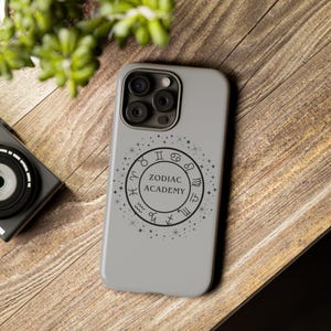 May include: A pale grey phone case with a black circular design featuring zodiac symbols and the words "ZODIAC ACADEMY". The case rests on a wooden surface, with a camera and foliage in the background.