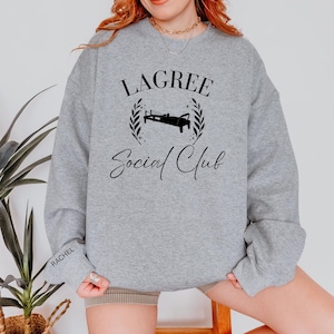 May include: A heather gray sweatshirt with the text "LAGREE Social Club" in black script. The design includes a black illustration of a Lagree fitness machine, surrounded by a laurel wreath. The name "RACHEL" is printed on the left sleeve.