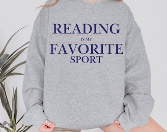Könnte beinhalten: Ein hellgraues Sweatshirt mit dem Text "READING IS MY FAVORITE SPORT" in Marineblau. Der Text ist mittig auf der Vorderseite des Sweatshirts platziert. Das Sweatshirt hat einen Rundhalsausschnitt und lange Ärmel.