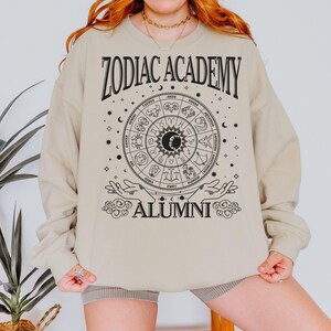 Op de afbeelding: Een beige sweatshirt met een zwarte afbeelding van een dierenriemwiel en de tekst "Zodiac Academy Alumni".