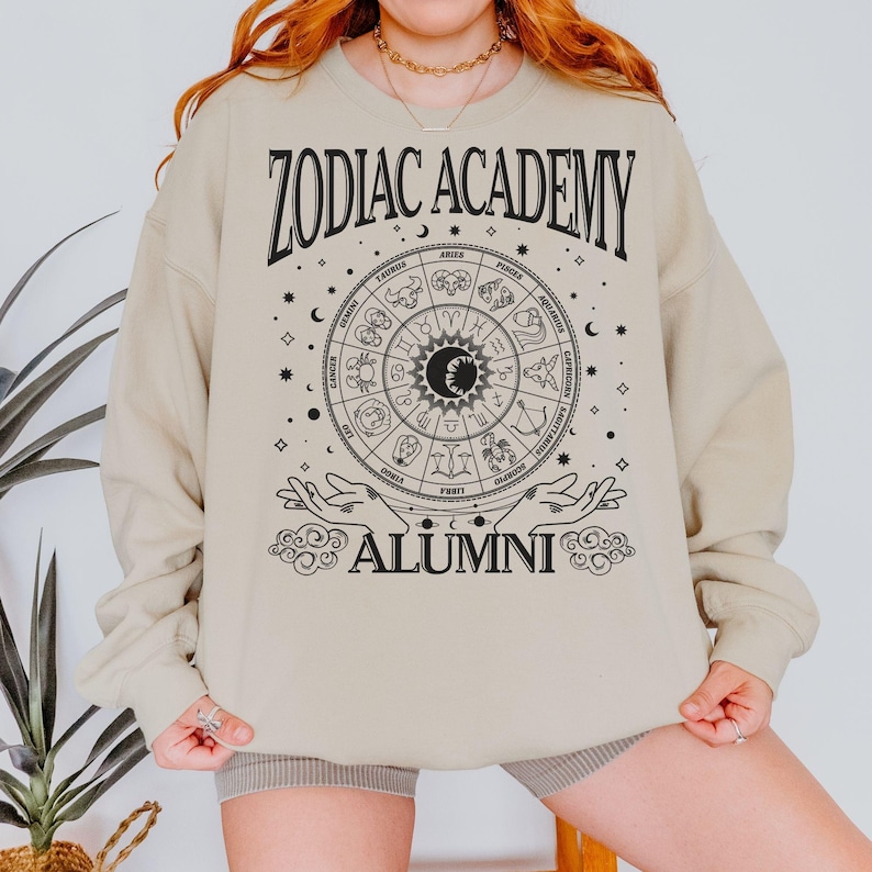 Op de afbeelding: Een beige sweatshirt met een zwarte afbeelding van een dierenriemwiel en de tekst "Zodiac Academy Alumni".
