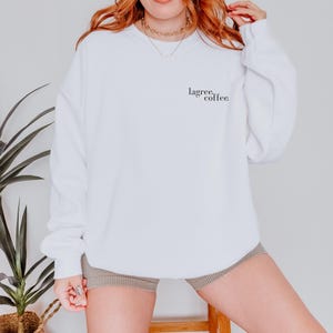 May include: A white sweatshirt with the text "lagree, coffee" in black script on the upper left chest. The sweatshirt has long sleeves and a crew neck. The model is wearing tan shorts and a gold necklace.