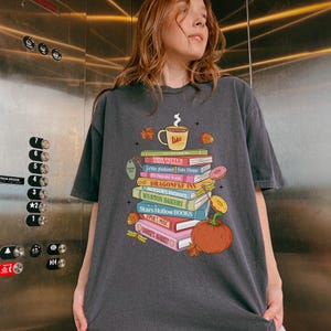 Puede incluir: Camiseta gris oscuro con una pila de libros, incluyendo títulos como "MISS PATTYS" y "DRAGONFLY INN". También se muestra una taza de café con "Lukes" y una calabaza. El diseño tiene un tema otoñal.