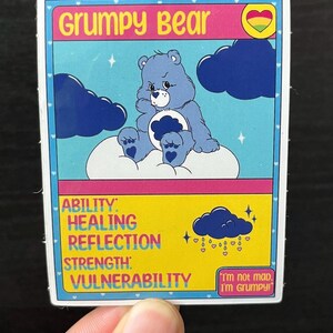 May include: A sticker featuring Grumpy Bear from the Care Bears. The blue bear sits on a cloud, surrounded by dark blue clouds and stars. Text includes "Grumpy Bear," and "I'm not mad, I'm Grumpy!" Additional text lists abilities.