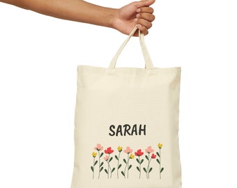 May include: A beige canvas tote bag with the name "SARAH" printed in black. The bag has a row of colorful flowers printed at the bottom.