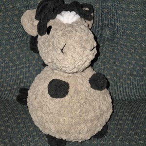 May include: A handmade plush toy in the shape of a cow. The toy is primarily light gray with black accents, including spots, ears, and a tuft of hair. The eyes are stitched with black thread.