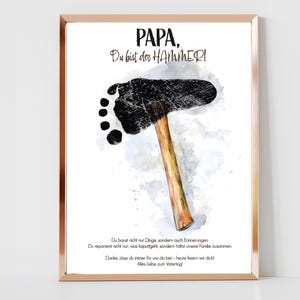 Puede incluir: Una ilustración de acuarela de un martillo con una huella de pie negra en la cabeza del martillo. El texto "PAPA, Du bist der HAMMER!" está escrito en negro en la parte superior de la imagen. El texto debajo de la imagen dice "Du baust nicht nur Dinge, sondern auch Erinnerungen. Du reparierst nicht nur, was kaputtgeht, sondern hältst unsere Familie zusammen. Danke, dass du immer für uns da bist - heute feiern wir dich. Alles Liebe zum Vatertag."