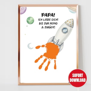 Pode incluir: Impressão emoldurada com um foguete branco e uma impressão de mão laranja. O texto diz 'PAPA! ICH LIEBE DICH! BIS ZUM MOND & ZURÜCK!'. Planetas e uma lua crescente decoram o fundo. Um círculo vermelho com 'SOFORT DOWNLOAD' está no canto inferior direito.