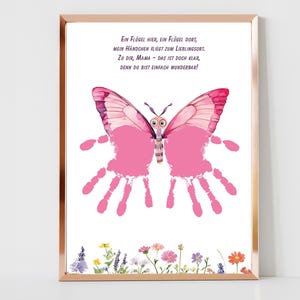 Puede incluir: Impresión enmarcada con un diseño de mariposa rosa, alas de huellas de manos y un borde floral. La mariposa tiene un efecto acuarela. Texto en alemán en la parte superior.