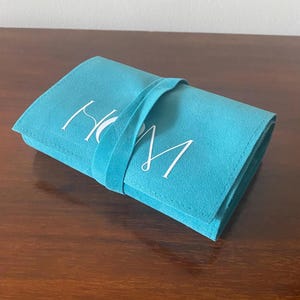 以下が含まれることがあります： ターコイズブルーのスエード調の折りたたみ式ケース。お揃いのストラップ留めが付いています。ケースの前面には、白い筆記体で「H」と「M」の文字が書かれています。ケースは濃い茶色の木製の表面に置かれています。