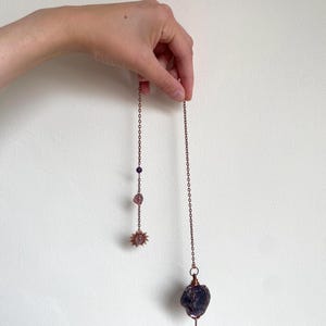 May include: Two copper-toned pendulums hang against a white backdrop. One has a sun-shaped charm and amethyst beads. The other has a large, dark purple crystal at the end. The pendulums are held by a hand.