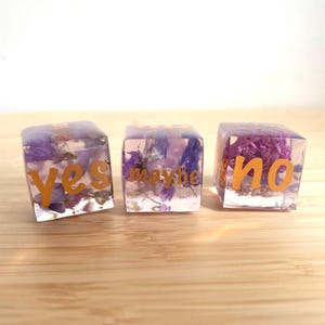 Peut inclure: Trois cubes de résine transparente avec des fleurs violettes séchées à l'intérieur. Les cubes portent les mots "yes", "maybe" et "no" en lettres orange. Posés sur une surface en bois brun clair.