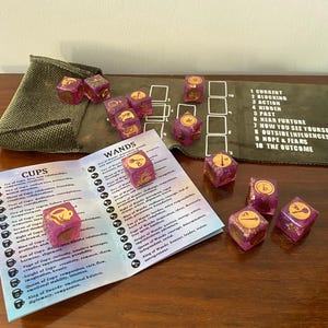Peut inclure: Un ensemble de dés violets et dorés avec des symboles de tarot, disposés sur un tapis vert avec des cases numérotées et une carte listant les significations du tarot. Une petite pochette verte est également présente. Accessoire de jeu unique.