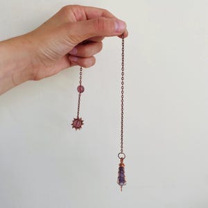 Puede incluir: Un péndulo de color cobre con un cristal púrpura al final de una cadena. El péndulo está sostenido por una mano. Una cadena más pequeña con una cuenta rosa y un dije en forma de sol cuelga a la izquierda.