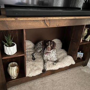 May include: A dark brown wooden entertainment center with a built-in dog bed. The dog bed is lined with a white fluffy blanket. A brown and white dog is lying on the bed. The entertainment center has shelves on the sides and a shelf above the dog bed.
