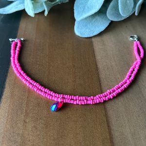 May include: A pink beaded bracelet with a pink crystal charm. The bracelet is made of two strands of beads and has a silver clasp.