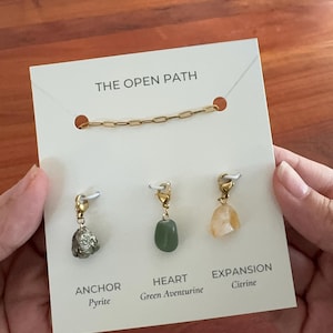 May include: A gold chain necklace and three gemstone charms on a white display card labeled THE OPEN PATH. The charms include a metallic pyrite labeled ANCHOR, a smooth green aventurine labeled HEART, and a translucent citrine labeled EXPANSION.