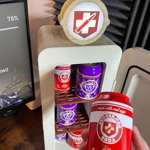 May include: A small, off-white mini-fridge with a red and white JUGGER-NOG SODA can in hand. The fridge is stocked with red and purple soda cans. The top of the fridge has a circular emblem with a red cross and bullet. The can reads "made with real eggs".