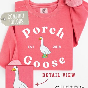 May include: Watermelon-colored custom sweatshirt with the words "Porch Goose" and a goose graphic. The sweatshirt also has the words "Comfort Colors" and "Custom Sweatshirt" on it.