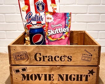 May include: A wooden crate with the text Grace's MOVIE NIGHT, featuring a ticket, clapperboard, 3D glasses, and star icons. Inside are a bag of Butter popcorn, a bag of Desserts Skittles, and a blue Pepsi can, all arranged for a movie night.