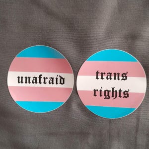 Könnte beinhalten: Zwei runde Aufkleber mit dem Transgender-Flaggen-Design. Einer trägt den Schriftzug "unafraid" in gotischer Schrift. Der andere trägt den Schriftzug "trans rights" in gotischer Schrift. Die Aufkleber sind blau, rosa und weiß.