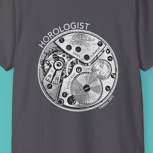 Może przedstawiać: Szary t-shirt z białym nadrukiem mechanizmu zegarka i napisami "HOROLOGIST" i "OMEGA".