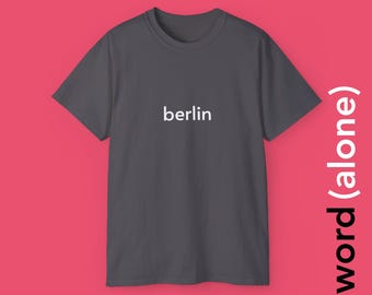 May include: A dark gray t-shirt with the white word "berlin" printed across the chest. The shirt is displayed against a pink background with the words "word (alone)" in white on the right side.